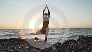 Flexible caucasian girl doing tree yoga asana at seashore at sunset.