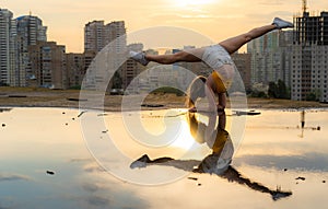 Flexible female gymnast doing handstand and calisthenic with reflection in the water on cityscape background during