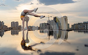 Flexible female gymnast doing handstand and calisthenic with reflection in the water on cityscape background during