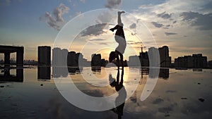 Flexible female gymnast doing handstand and calisthenic with reflection in the water on cityscape background during