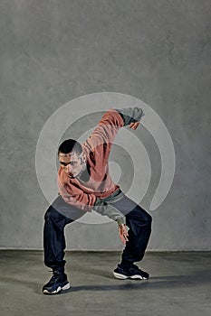 Flexible male with tattooed face, beard. Dressed in colorful jumper, black pants and sneakers. Dancing on gray