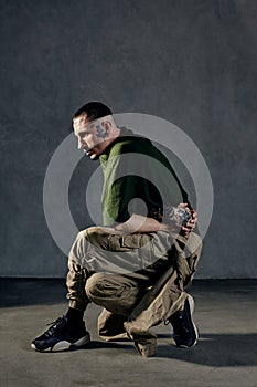 Flexible man with tattooed body, earrings, beard. Dressed in khaki t-shirt, overalls, black sneakers. Dancing on gray