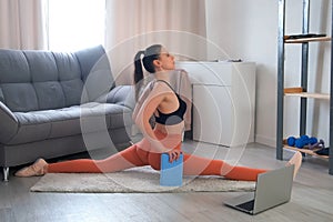 Flexible sporty young woman making longitudinal split sitting on carpet at home.