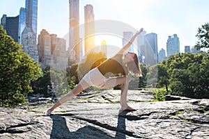 Flexible woman doing yoga pose in city park in New York at sunset time.