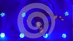 Flexible woman hanging in an aerial acrobatics ring performs complex tricks. Aerial acrobat in a dark studio with blue