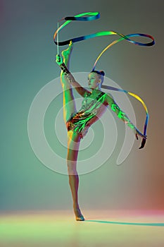 Flexible young girl, female rhythmic gymnast training with ribbon isolated over green background in neon light