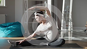 Flexible young pregnant woman bending aside sitting on exercise mat on sunny morning at home. Wide shot portrait of fit