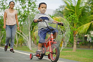 Flifestyle portrait of Asian Indonesian mother and young happy son at city park having fun together the kid learning bike riding