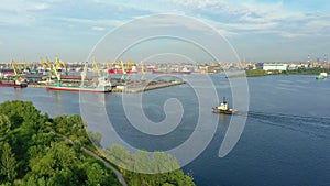 Flight in from the air over the cargo port of a major city, over green vegetation trees. Tugboat ship sails in the bay of the bay,