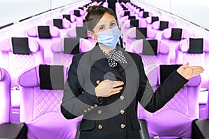 A flight attendant with a surgical mask points with her arms in the aisle of an airplane. Subject on a blurred background