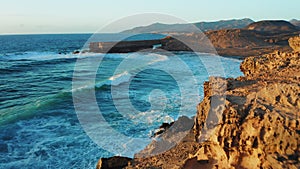 Flight over wide ocean waves crashing among huge rock formations. Sunset light. Turquoise water foaming. Aerial