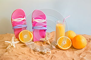 Flip flops, orange juice, oranges, starfishes and seashells on sand beach on blue backdrop.