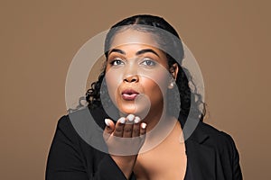 Flirt concept. Cheerful brazilian body positive woman sending air kisses to camera, posing over brown background