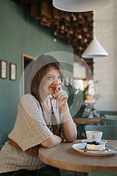Flirtatious girl sits at table in cafe. Young woman in cafe with cup of latte or cappuccino and dessert