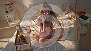 Flirting girl talking mobile phone in sunny bedroom. Smiling woman resting bed