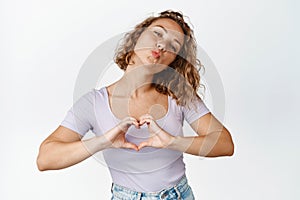 Flirty blond girl pucker lips, kissing and showing heart gesture, shows affection, stands against white background