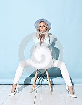 Flirty gorgeous blonde curly woman in white blue pantsuit and high-heeled shoes sitting backwards on stylish white chair