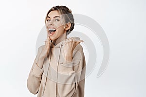 Flirty young woman pointing finger right and laughing coquettish, standing in beige hoodie against white background