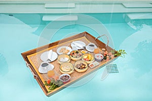 Floating Breakfast tray in swimming pool at luxury hotel or tropical resort villa, fruits, croissant, coffee, and orange juice.