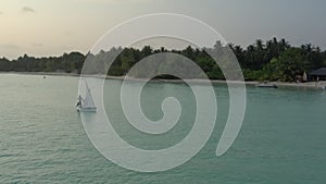 Floating small sailboat with sails up in calm sea anchored near tropical islands in Maldives