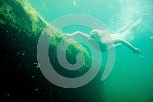 A floating woman. Underwater portrait. Girl in white dress swimming in the lake. Green marine plants, water