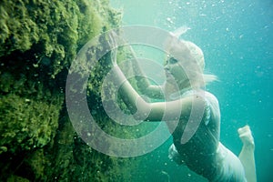 A floating woman. Underwater portrait. Girl in white dress swimming in the lake. Green marine plants, water