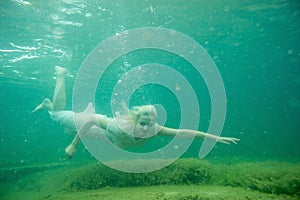 A floating woman. Underwater portrait. Girl in white dress swimming in the lake. Green marine plants, water