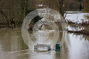 Flooded parkland with a debris coated bench and litter bin.