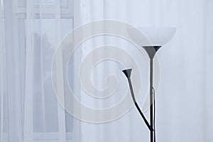 Floor electric lamp in black with white lampshades in the interior of the room against background of white curtains and windows