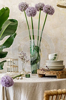 Floral composition at kitchen interior with wooden family table, beautiful flowers in vase, plates, cups, tray .