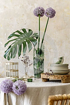 Floral composition at kitchen interior with wooden family table, beautiful flowers in vase, plates, cups, tray.