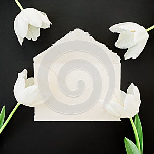 Floral composition with paper envelope and white tulips on black background. Flat lay, top view. Valentines day composition