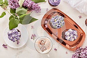 Floral purple cupcakes on the golden tray using trend Dreamy Escapism. Desserts, coffee cup and lilac flowers background