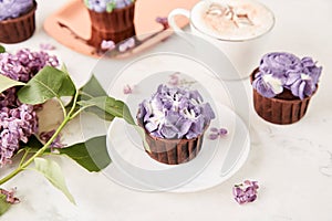 Floral purple cupcakes using trend Dreamy Escapism. Aesthetics desserts, coffee cup and lilac flowers background.