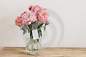 Floral still life scene. Pink peonies flowers, bouquet in glass vase on wooden table. Blank gift tag, label mockup