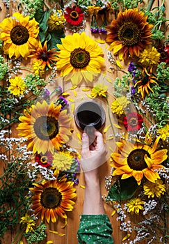 Floral summer background. A mug of coffee in a woman`s hand on a wooden background with sunflowers and wildflowers