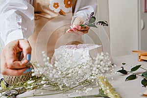 A florist decorates a gift box with flowers and a ribbon on a white desktop. Only the hands are in the frame