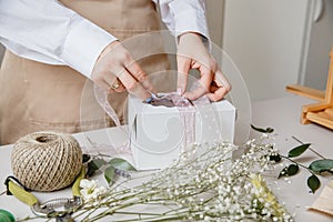 A florist decorates a gift box with flowers and a ribbon on a white desktop. Only the hands are in the frame