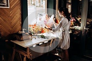 Florist dressed in a striped dress stands near the table with colorful dried flowers and holds transparent packaging