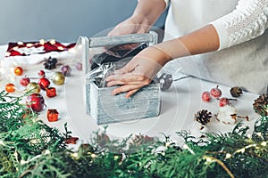 Florist woman makes Christmas composition for table decoration from thuja, spruce branches in wooden box. Using of floral foam,