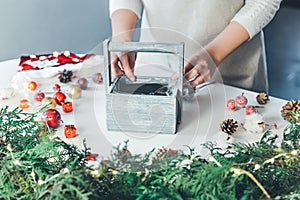 Florist woman makes Christmas composition for table decoration from thuja, spruce branches in wooden box. Using of floral foam,