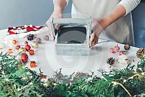 Florist woman makes Christmas composition for table decoration from thuja, spruce branches in wooden box. Using of floral foam,