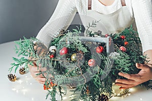 Florist woman makes Christmas composition for table decoration from thuja, spruce branches in wooden box. Using of floral foam,