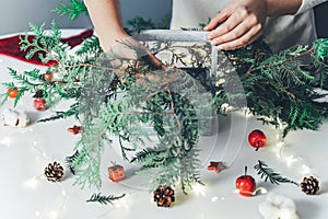 Florist woman makes Christmas composition for table decoration from thuja, spruce branches in wooden box. Using of floral foam,