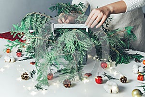 Florist woman makes Christmas composition for table decoration from thuja, spruce branches in wooden box. Using of floral foam,