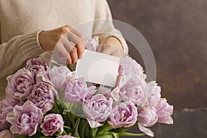 Florist at work: pretty young woman making a big bouquet of double violet tulip flowers put a greeting card on a dark rustik