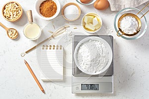 Flour in bowl on digital scale with notepad and pen and baking ingredients on marble table