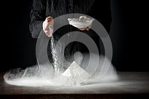 Flour on a kitchen table on a black moody background in the morning light. Woman`s hands sieving flour through a sieve.