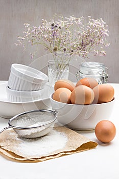 Flour and sieve on paper. The egg is on table. Brown chicken eggs in bowl. White ceramic dishes