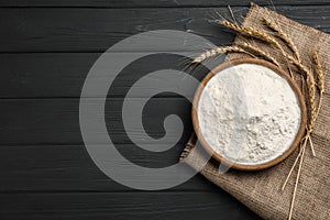 Flour, wheat ears and sackcloth on black wooden table, flat lay. Space for text